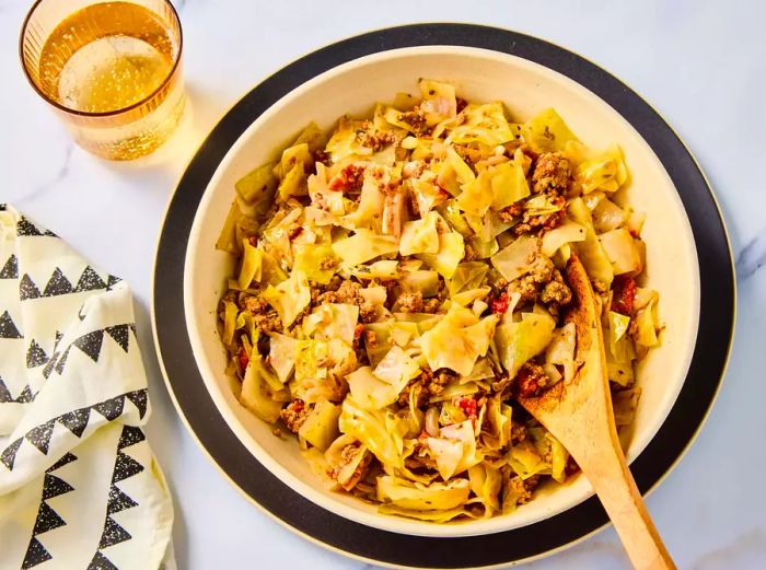 A bowl filled with ground beef and cabbage, accompanied by a wooden serving spoon and a glass of sparkling water