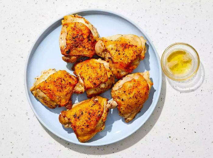 A top-down view of cooked chicken thighs served on a blue round plate, accompanied by reserved drippings in a small bowl.