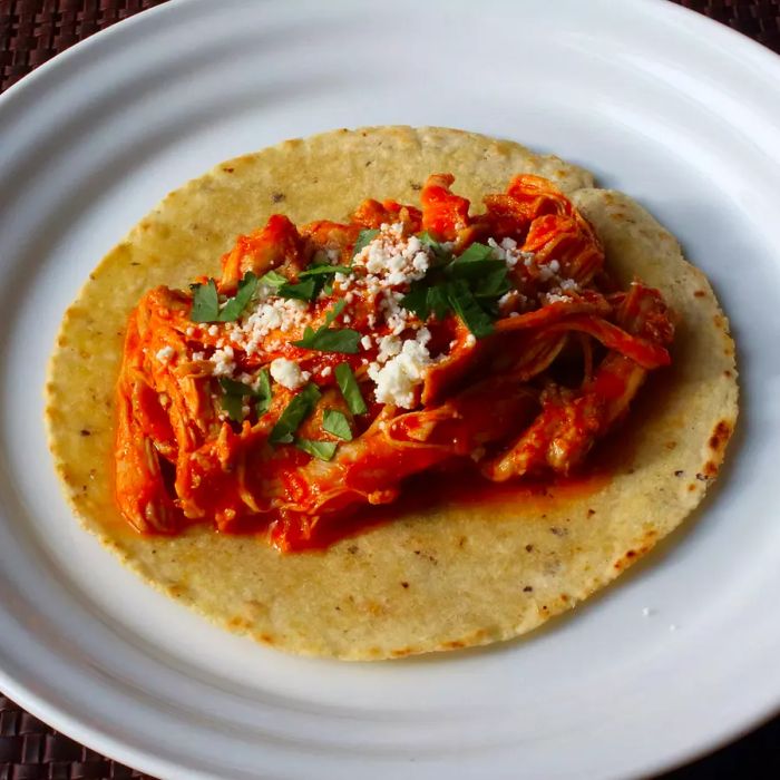 A close-up of a Chicken Tinga taco, topped with cheese and fresh herbs, served on a white plate.