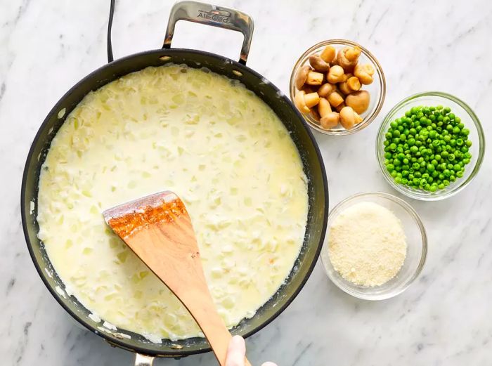 A top-down view of milk being poured into the skillet with onions, alongside a small bowl containing mushrooms, peas, and cheese.