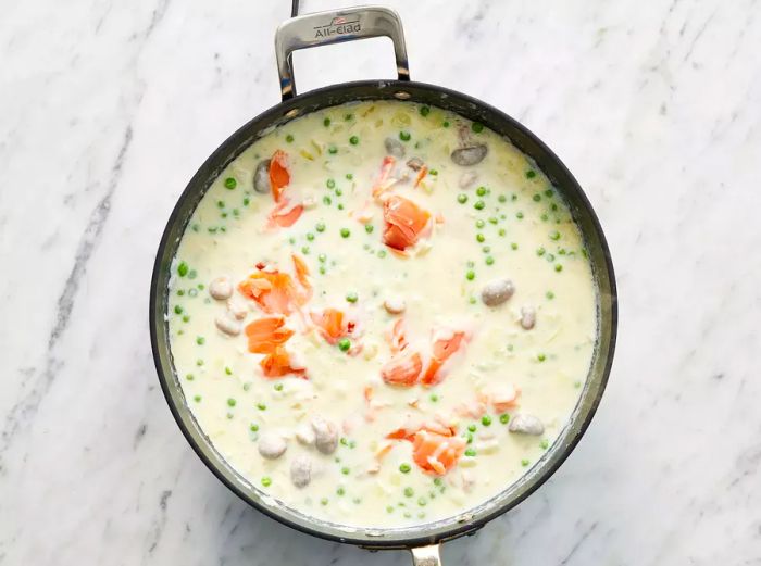 A top-down view of cheese, peas, and smoked salmon being added to the creamy sauce