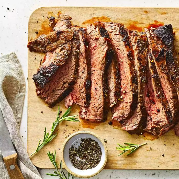 Sliced broiled balsamic-soy flank steak served on a wooden board, accompanied by a bowl of freshly cracked black pepper and sprigs of fresh rosemary.