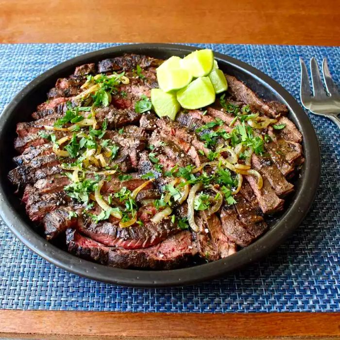 A close-up shot of tender slices of Grilled Mojo Beef, served with caramelized onions, fresh herbs, and wedges of lime on the side.