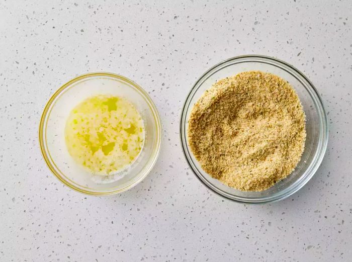 Melted butter in a glass dish placed beside the breadcrumbs in a separate glass bowl.