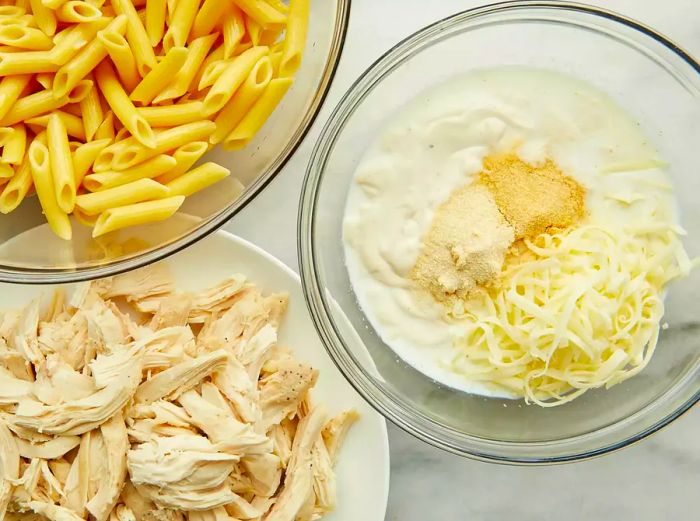 Aerial view of pasta, chicken, and an unmixed Alfredo mixture in separate bowls.