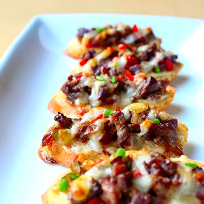 Close-up view of Mini Philly Cheesesteaks on bread slices, garnished with green onions, on a white platter.