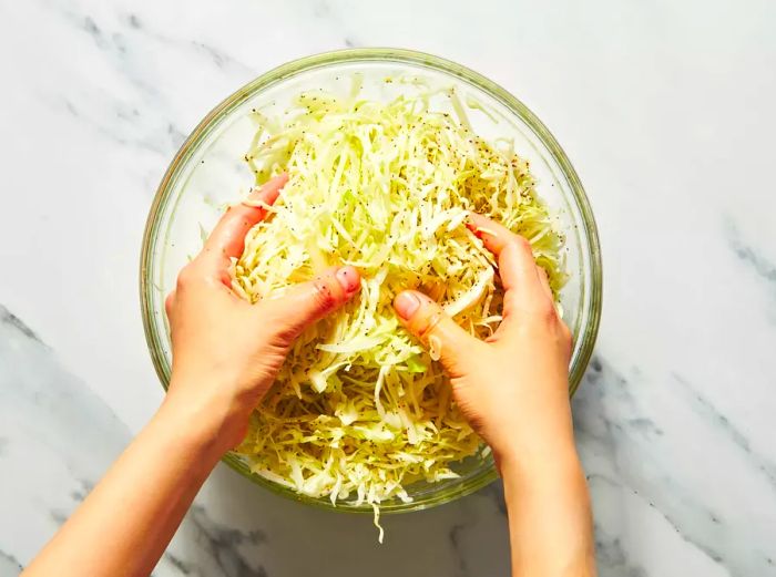 Hands mixing shredded cabbage in a glass bowl for slaw preparation.