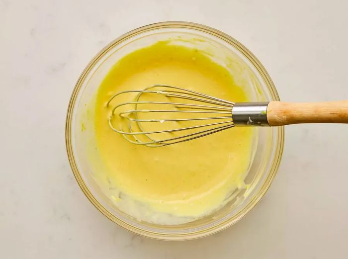 A large glass bowl filled with mayonnaise, honey, mustards, and lemon juice being whisked together.