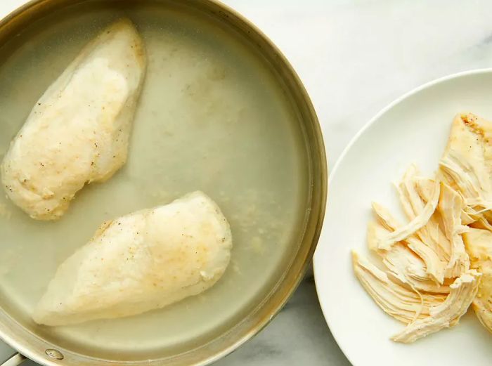 Aerial view of chicken breasts cooking in a skillet with water, and chopped chicken pieces plated on the side.