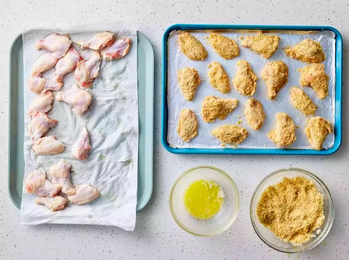 Chicken wings placed on a baking sheet next to the breading station, with breaded wings set aside on a separate baking sheet.