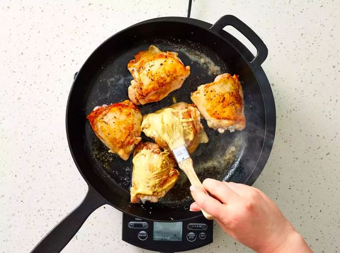 Applying Dijon mustard by hand to chicken thighs in a sizzling cast-iron skillet on the stove