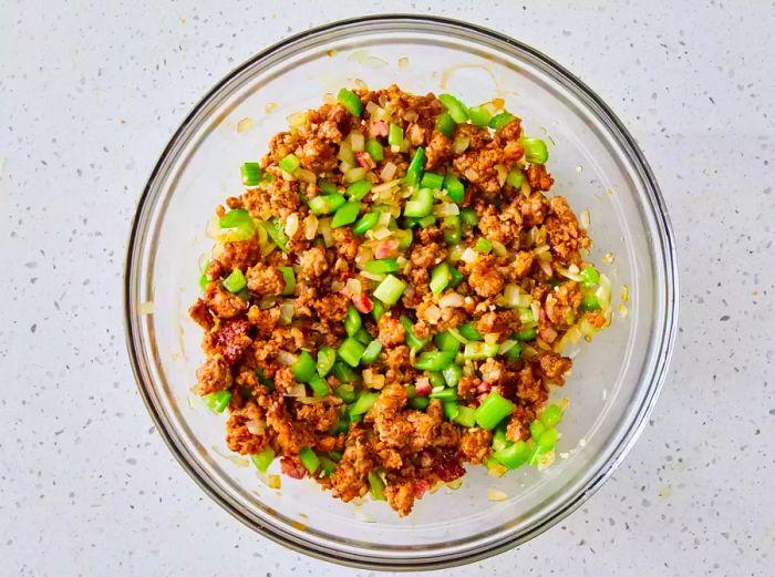 A glass bowl filled with cooked ground sausage, pancetta, onion, garlic, and celery