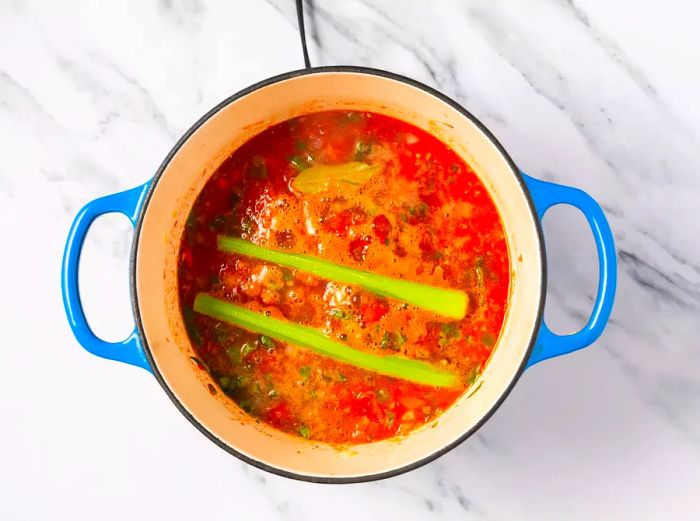 A large pot filled with chopped vegetables in pureed tomato sauce, with two stalks of celery and a bay leaf