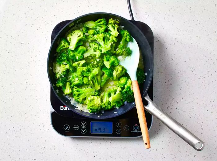 A skillet with broccoli and chopped onions being stirred using a silicone spatula