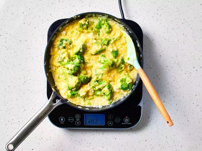 A skillet with rice, cheese sauce, and condensed soup mixed into the broccoli and onions