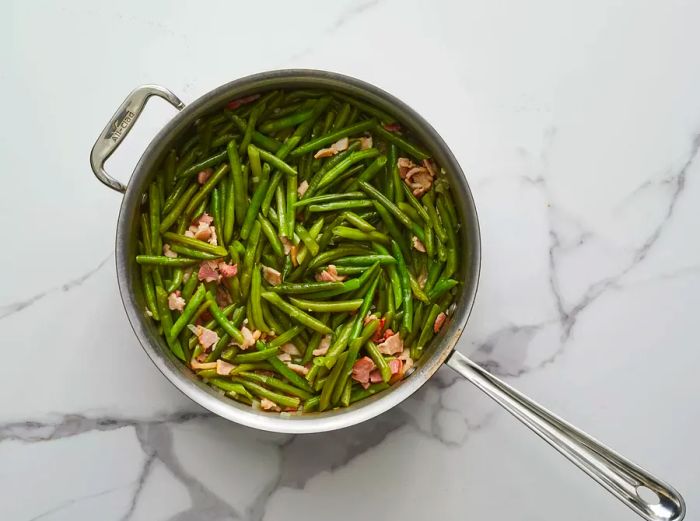 An overhead shot of green beans cooking in a skillet with bacon, onions, and garlic.