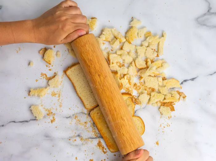 A top-down view of a rolling pin crushing dried bread slices, preparing them for stuffing.