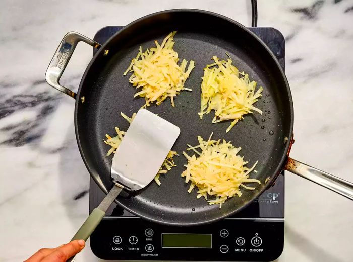 An oiled frying pan with four German potato pancakes, one being gently pressed with a metal spatula