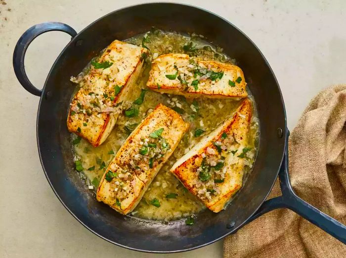 A close-up view of a skillet with halibut fillets, topped with a wine sauce and garnished with freshly chopped parsley.