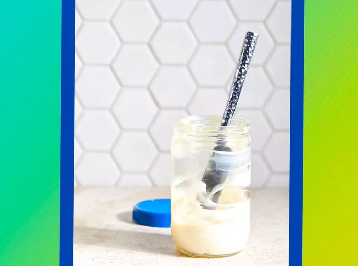 A jar of mayonnaise with a knife sticking out, sitting on a counter with the lid nearby, against a blue-to-green ombre backdrop