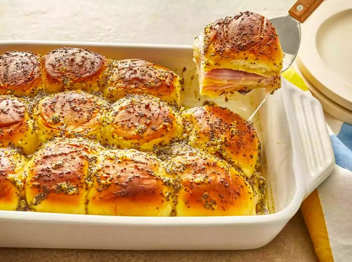 A single ham and cheese slider being lifted from a casserole dish filled with delicious sliders.