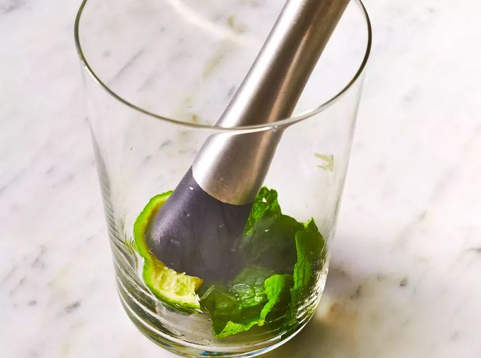 A clear glass with mint leaves and a lime wedge being muddled together.