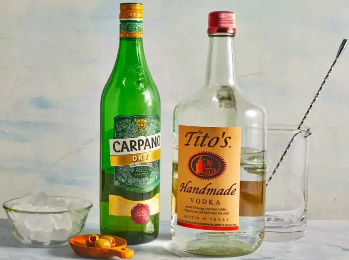 Bottle of vermouth, bottle of vodka, small bowl of Spanish olives, a pitcher with a stirring spoon, and a bowl of ice.