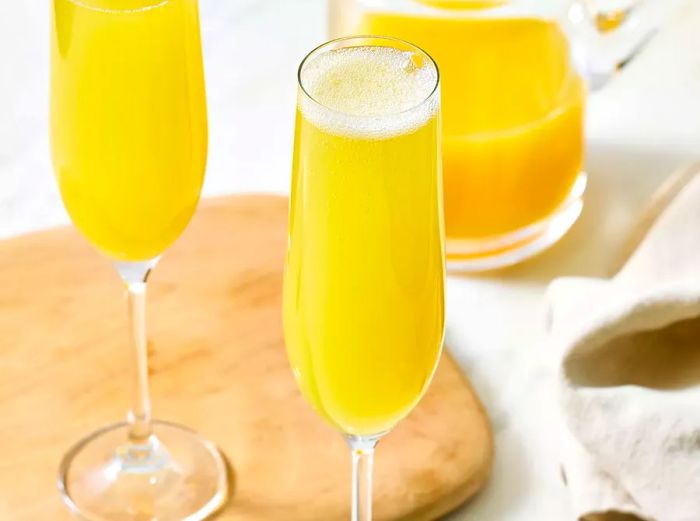 Two glasses of mimosa placed alongside a pitcher of orange juice on a wooden board.