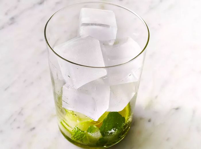 A clear glass with muddled mint, lime, and sugar, now filled with ice.