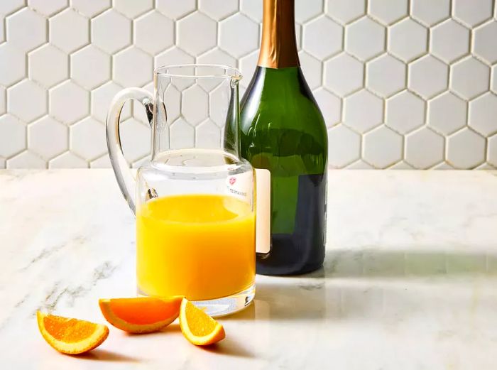 A pitcher of orange juice alongside a bottle of Champagne and slices of orange.