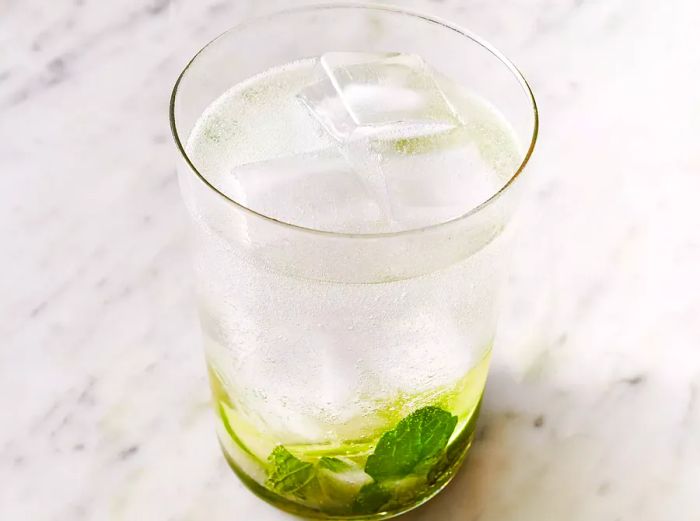 A high-angle shot of a clear glass with rum and ice poured over muddled sugar and lime.