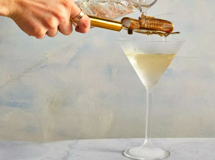 A hand pouring from a pitcher through a cocktail strainer into a chilled martini glass.