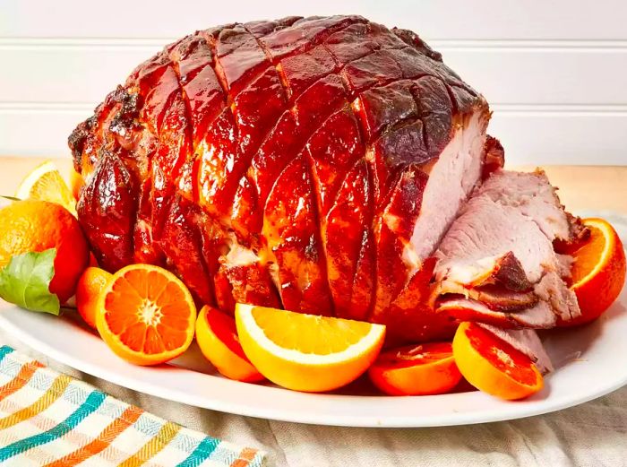 Close-up shot of Tangy Honey-Glazed Ham sliced on one side, served on a platter with citrus fruits.