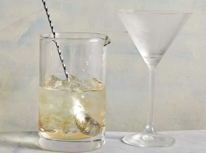 A small glass pitcher filled with a light amber liquid on ice, accompanied by a stirring spoon inside and a frosted martini glass beside it.