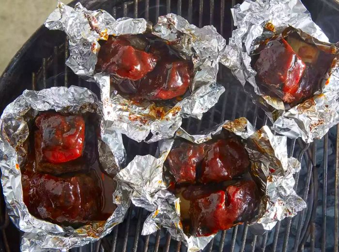 Aerial view of ribs in tin foil on the grill, being brushed with barbecue sauce.