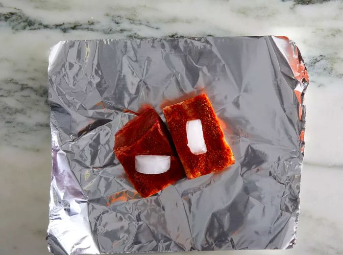 An overhead shot of two ribs placed in the center of a sheet of tin foil, with one ice cube on top.