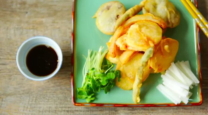 A plate of crispy vegetable tempura served with soy sauce and chopsticks.