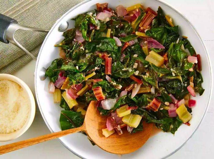 A top-down view of a large skillet filled with sautéed Swiss chard and parmesan cheese, with a spoonful resting in a bowl.