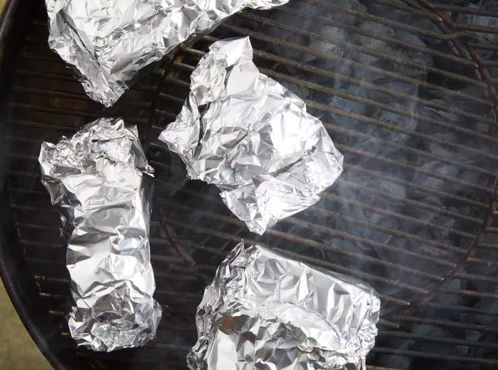 An overhead view of four foil-wrapped ribs cooking on a grill.