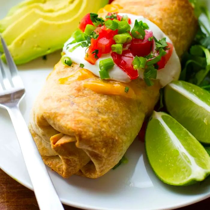 Baked Chicken and Rice Chimichanga served on a white plate, accompanied by a fork, lime wedges, and slices of fresh avocado.