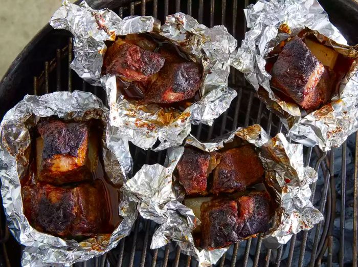 An aerial shot of opened foil packets with ribs inside, continuing to cook on the grill.