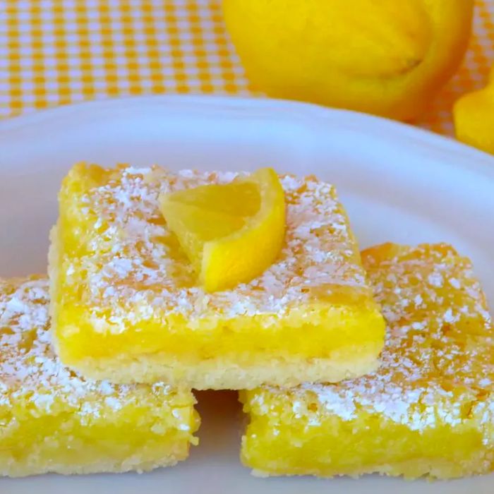 three lemon bars dusted with powdered sugar, stacked on a white plate, with whole lemons in the background