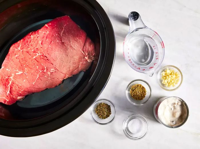 Aerial view of a London broil in a slow cooker surrounded by small bowls of condensed soup, salt, pepper, garlic, oregano, and basil.