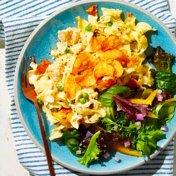 A comforting tuna noodle casserole topped with crispy crushed potato chips, served alongside a fresh salad on a turquoise plate.