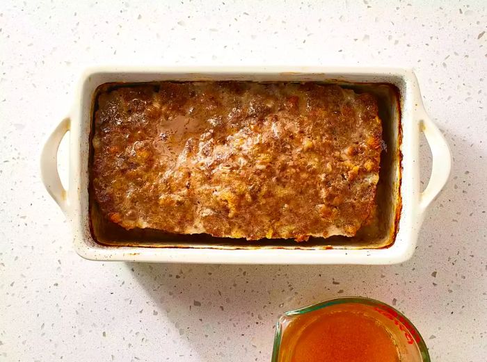 The baked meatloaf in the loaf pan, with the rendered fat collected in a measuring cup