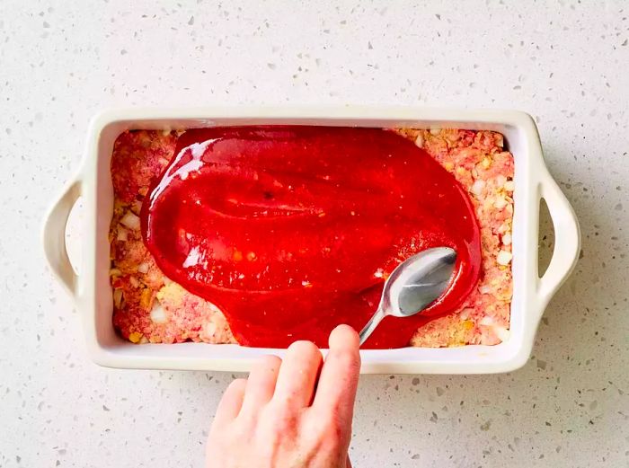 A loaf pan with brown sugar meatloaf being coated in a rich brown sugar-ketchup glaze.