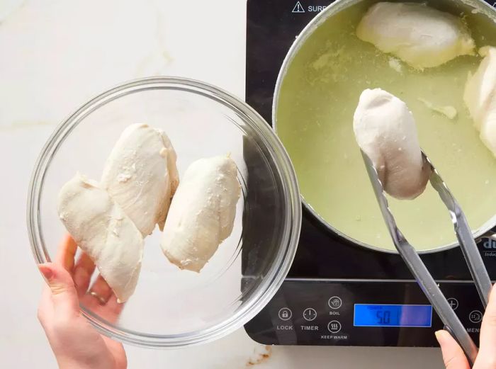 Someone lifting a cooked piece of chicken from a pot of water, with a bowl of chicken beside them