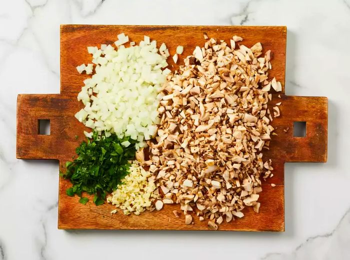 An overhead shot of mushrooms, onions, garlic, and fresh parsley chopped on a wooden cutting board.