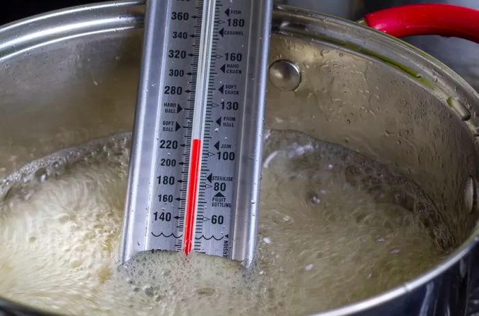 Candy thermometer placed in a modern metal pot with red handles, boiling apple jelly