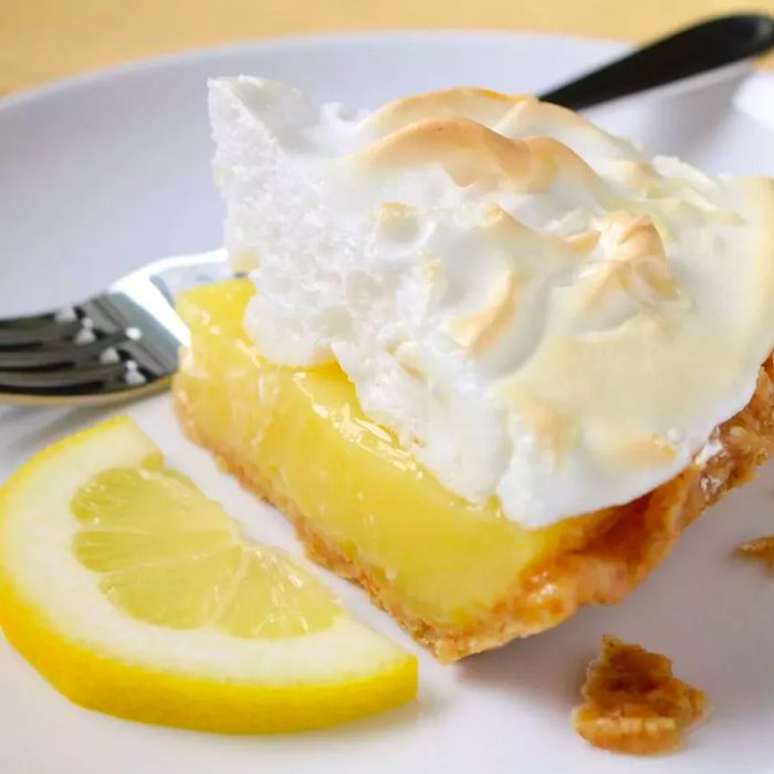 A slice of pie topped with golden-brown meringue, garnished with a lemon wedge, served on a pristine white plate.